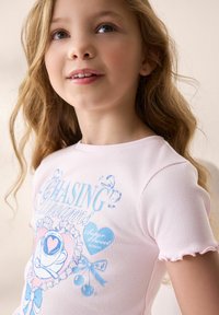 Lyserød ribbet T-shirt med korte, flagrende ærmer; har et blåt grafisk motiv af et hjerte og roser sammen med teksten "Chasing Dreams."