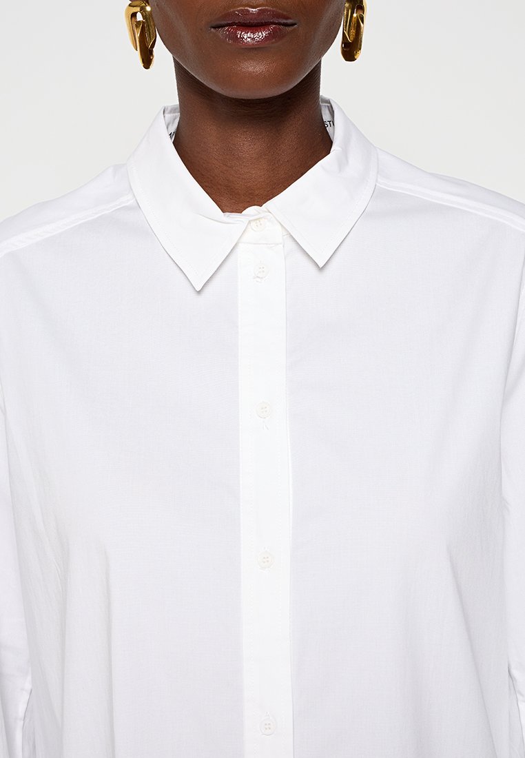 Donkerhuidige persoon die een nette witte overhemd met puntkraag en grote gouden oorringen draagt, op een neutrale achtergrond.