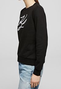 Sudadera negra con letras blancas bordadas en el frontal. Presenta un cuello redondo y puños acanalados, combinada con vaqueros de mezclilla azul claro.