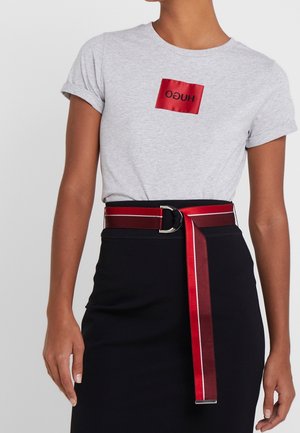 Femme portant un t-shirt gris clair avec un logo carré rouge, une jupe noire taille haute et une ceinture rouge avec une boucle argentée et un détail rayé.