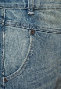 Jean en denim bleu clair avec une poche avant arrondie dotée d'un bouton en métal brun. Le tissu présente une texture légèrement décolorée.