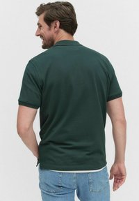 Mörkgrön polo med kort ärm, tillverkad av slät tyg. Har en klassisk krage och sprund på sidan av nederdelen, bärs tillsammans med ljusblå jeans.