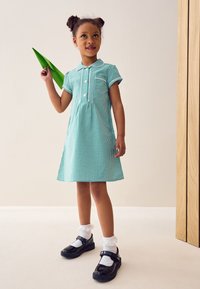 Grøn gingham-kjole med korte ærmer, krave og knapstolpe; kombineret med sorte lakskindssko og hvide flæse-sokker. Holder et grønt papirfly.