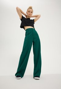 Jeune femme posant les mains derrière la tête, portant un haut court noir, un pantalon vert taille haute à jambes larges, et des baskets noires.