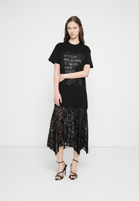 Čierne tričko s dizajnom z flitrového textu, vrstvené nad čiernou čipkovanou sukňou s vysokým a nízkym okrajom, doplnené čiernymi sandálmi na remienky.