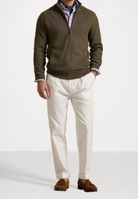 Muž v olivovém zipovém svetru, světle fialové košili, bílých kalhotách, hnědých mokasínech a námořnickém šátku s puntíky, stojící s rukama v kapsách.