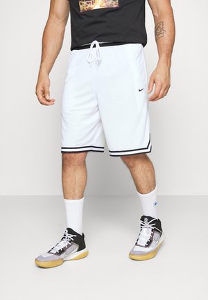 Homme portant un short de basketball Nike blanc avec un liseré noir, des chaussettes blanches Nike et des chaussures de basketball Nike noir et blanc, debout sur un sol uni.