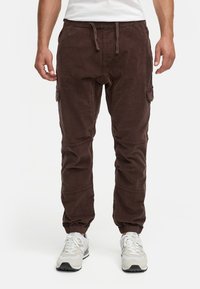 Pantalones de pana marrón con cintura elástica, cordón ajustable, tobillos con puños y bolsillos laterales tipo cargo. Combinados con zapatillas blancas.