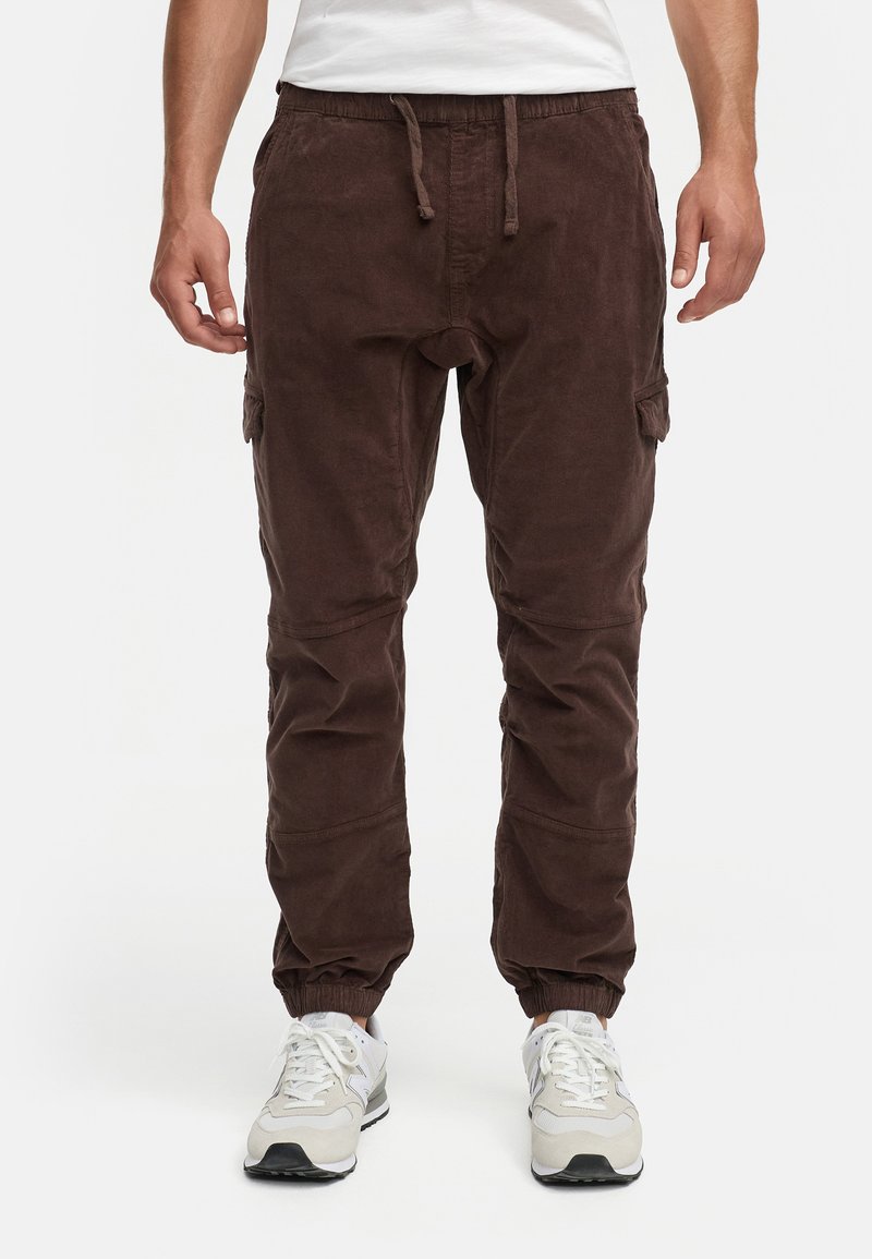 Pantalones de pana marrón con cintura elástica, cordón ajustable, tobillos con puños y bolsillos laterales tipo cargo. Combinados con zapatillas blancas.