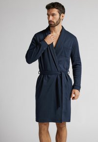 Tmavě modrý župan z měkké tkaniny, s šálovým límcem, dlouhými rukávy, náprsní kapsou a páskem v pase pro nastavitelnou velikost.