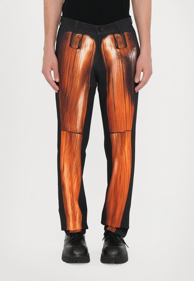 Schwarze Hose mit einem großen orangefarbenen Holzmaserungsmuster über den Beinen, subtilen gestickten Akzenten und einem Standardbund. Schwarze Schuhe.