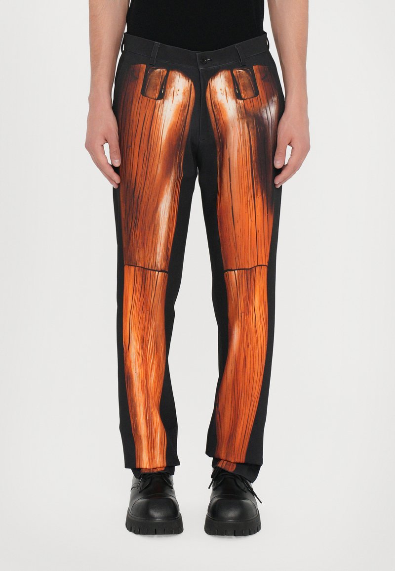 Schwarze Hose mit einem großen orangefarbenen Holzmaserungsmuster über den Beinen, subtilen gestickten Akzenten und einem Standardbund. Schwarze Schuhe.