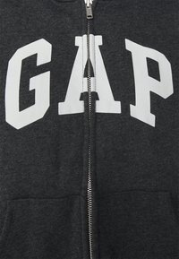 Felpa con zip di colore grigio antracite, caratterizzata da grande lettering bianco "GAP" sulla parte anteriore, texture liscia e zip metallica. Due tasche frontali.