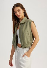 Gilet verde oliva corto con colletto, maniche corte, chiusura con zip, orlo elastico e una tasca laterale. Indossato sopra una maglietta bianca e pantaloni.