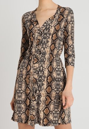 Femme portant une robe à imprimé serpent boutonnée avec des manches trois-quarts et un décolleté en V, sur un fond uni.