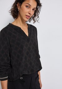Blouse noire avec un décolleté en V et des manches longues, présentant un motif géométrique en marron foncé. Le tissu a une texture lisse et une coupe décontractée.