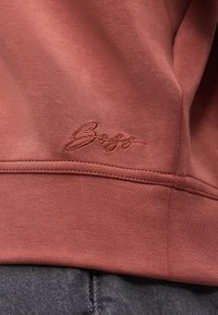 Ύφασμα σε αποχρώσεις σκουριάς με κεντημένο λογότυπο με καλλιγραφική γραφή κοντά σε ραφή ραμμένη οριζόντια πάνω από σκουρόχρωμα γκρι τζιν.