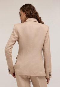 Beżowa marynarka o dopasowanym kroju, z wykładanym kołnierzem i trzema guzikami na rękawach. Gładka faktura materiału.