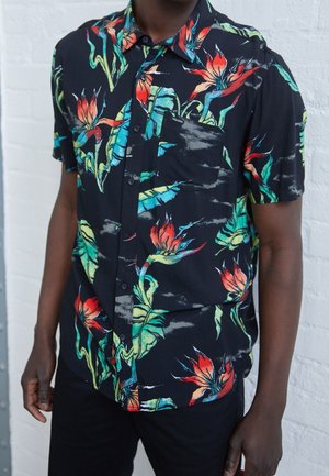Czarna koszula na krótki rękaw zapinana na guziki z żywym czerwono-zielonym tropikalnym kwiatowym wzorem, noszona przez osobę na tle jasnej, fakturowanej ściany.