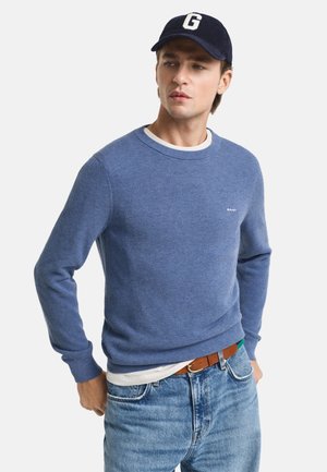 Blauwe gestructureerde trui met een ronde hals, gedragen over een wit shirt. Gecombineerd met lichte denim jeans en een donkerblauwe pet met een witte letter.