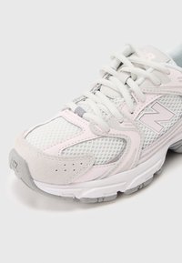 Valkoinen ja vaaleanpunainen lenkkikenkä, valmistettu verkkokankaasta ja mokkanahasta, jossa on kuvioitu pohja, näkyvät nauhat ja heijastava logo sivulla.