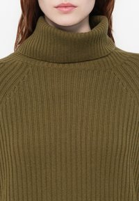 Olivno zelena rebrasta turtleneck pulover z visokim ovratnikom in teksturiranim pletenjem. Tkanina se zdi mehka s rahlim elastičnim učinkom.