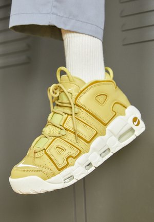 Pied portant une basket Nike Air More Uptempo jaune avec une semelle blanche et une chaussette blanche, sur fond de casier gris.