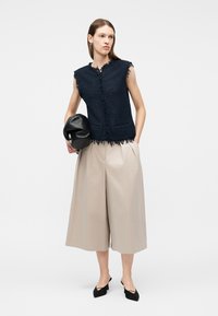Donna con top senza maniche testurizzato blu navy e pantaloni beige ampi e corti, che tiene una pochette nera e indossa tacchi neri.