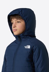 Laivastonsininen toppatakkimallinen takki hupulla, tikattu kuvio, vetoketjukiinnitys ja valkoinen logo vasemmassa rinnassa. Sileä pinta, kevyt ja istuva malli.