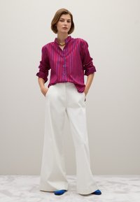 Vrouw in een rood en blauw verticaal gestreept shirt, brede witte broek, blauwe schoenen en een gouden kettinghang met haar handen in haar zakken.