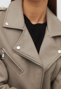 Beige faux leather jacket with silver hardware, zippered pocket, and wide collar. Layered over a black sweater. Smooth texture, modern design.