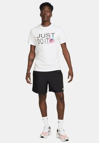 T-shirt bianco Nike con testo "JUST DO IT" e logo rosa, pantaloni corti neri, sneaker bianche e rosa e calze bianche con accenti neri del logo.