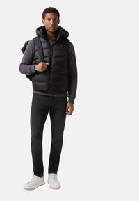 Zwarte gewatteerde vest met capuchon over een donker sweatshirt, gecombineerd met donker jeans en witte sneakers. Draagt een zwarte rugzak.