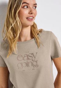 Camiseta de manga corta color beige con cuello redondo, que presenta un estampado gráfico texturizado que dice "THE easy going" en tonos marrones.