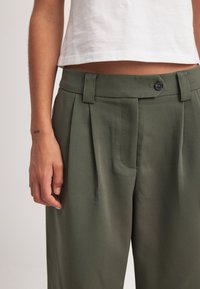 Khaki nabranne hlače s gladko, teksturirano površino, ki imajo zaponko na gumb in zanke za pas, v kombinaciji z belo kratko majico.