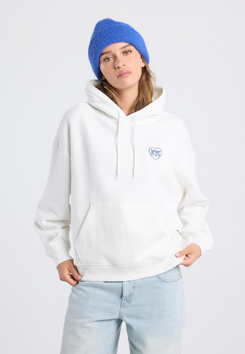 Young woman wearing a white NYC heart-printed hoodie, light blue jeans, and a blue knit beanie, standing against a plain light background.