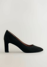 Zapato de tacón alto de ante negro con punta afilada y tacón cuadrado. El tacón presenta un sutil acento plateado. Textura suave en general.