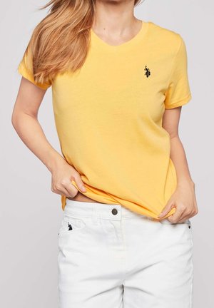 Vrouw die een gele korte mouw blouse en een witte broek draagt, met beide handen de zoom van het shirt ter hoogte van de taille vasthoudend.