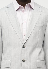 Veste de costume gris clair à fines rayures portée sur une chemise rose pâle boutonnée au col et au milieu, sur une personne à la peau foncée.