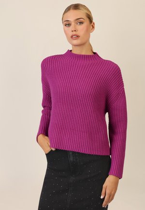 Lila gestrickter Pullover mit hohem Kragen, gerippter Textur und langen Ärmeln. Kombiniert mit einem schwarzen Jeansrock mit silbernen Nieten.