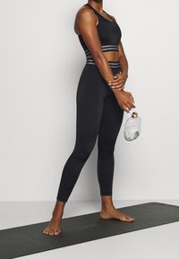 Mujer con ropa deportiva negra de pie descalza sobre una esterilla de yoga, sosteniendo una botella de agua transparente con una base de patrón de mármol.