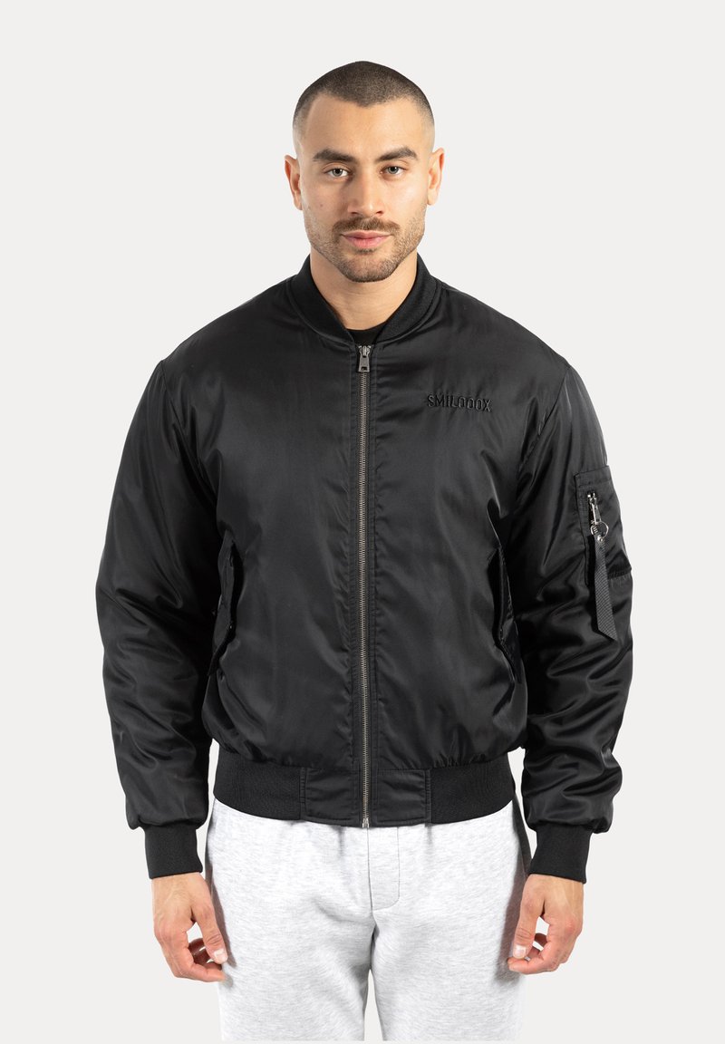 Giacca bomber nera realizzata in materiale liscio e lucido. Presenta una cerniera frontale, polsini a costine e una tasca con zip sulla manica sinistra.