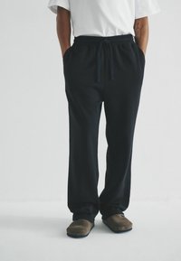 Schwarze Jogginghosen mit elastischem Bund und Kordelzug, seitlichen Taschen und bequemer Passform. Getragen mit braunen Slippers.
