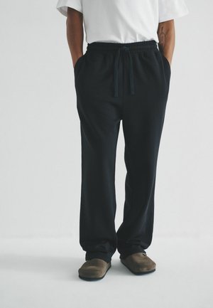 Schwarze Jogginghosen mit elastischem Bund und Kordelzug, seitlichen Taschen und bequemer Passform. Getragen mit braunen Slippers.