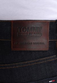 Darka denim jeans har en brun läderlapp med "TOMMY JEANS" präglat och "AN AMERICAN ORIGINAL" nedan, accentuerat av orange sömnad.