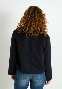 Veste en mélange de laine bleu marine avec un design structuré, dotée de manches longues, d'une texture légère et de coutures au dos. Associée à un jean en denim bleu.