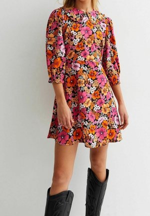 Woman wearing a short floral dress with pink, orange, and white flowers and black knee-high boots, standing against a light background.