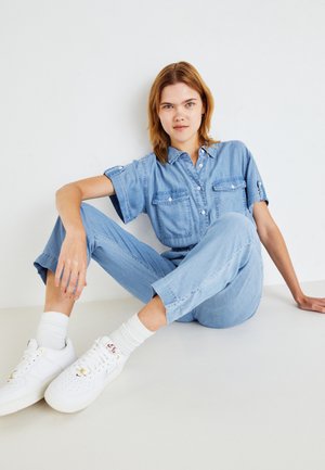 Jeune personne avec des cheveux blonds assise sur le sol, portant une combinaison en denim bleu clair et des baskets blanches sur fond blanc.