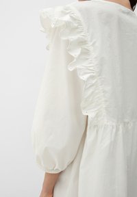 Weißes Kleid mit einem rüschelnen Schulterdesign und Ausschnitten, Ballonärmeln mit elastischen Bündchen und einem gerafften Saum. Aus weichem, leichtem Stoff.