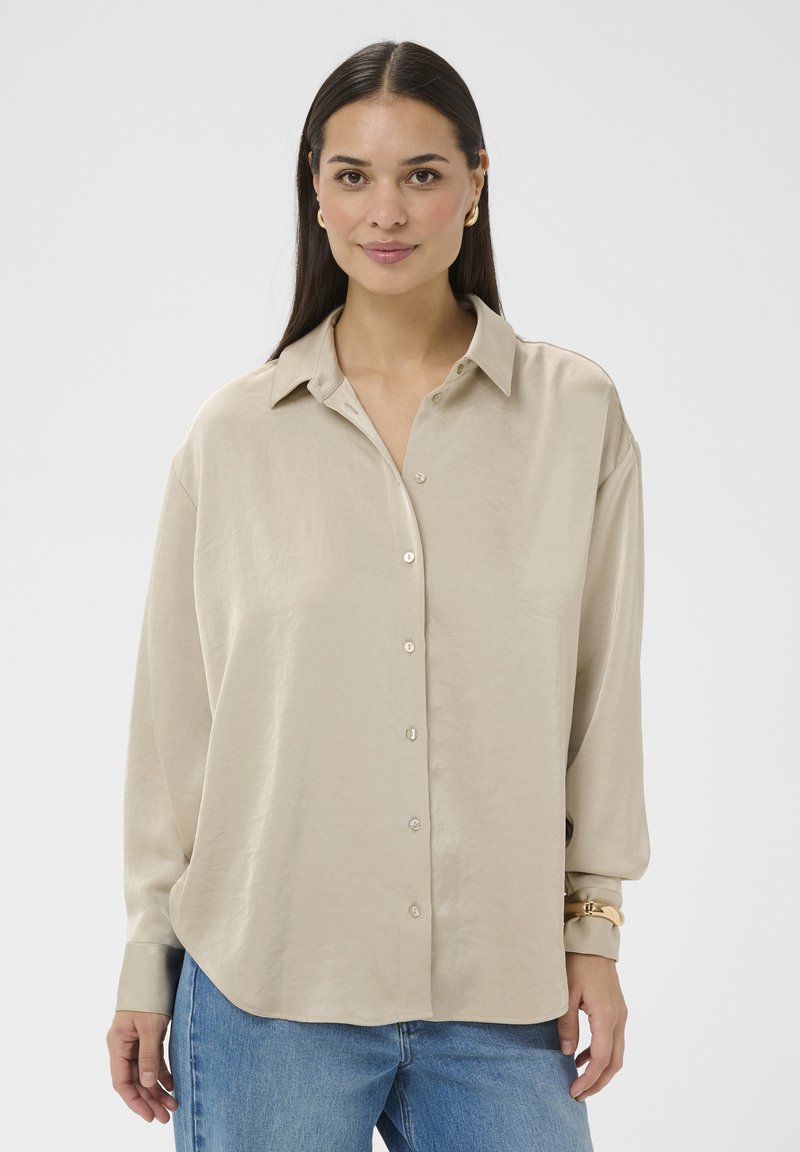 Femme aux longs cheveux foncés portant une chemise beige boutonnée, un jean bleu, des boucles d'oreilles créoles dorées et un bracelet en or, sur un fond blanc uni.
